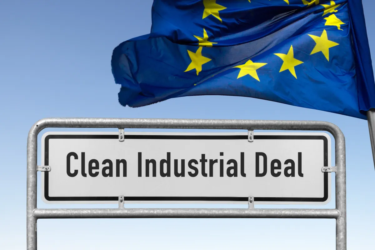 Blick auf ein weißes Ortsangabenschild mit der Aufschrift "Clean Industrial Deal" - daneben weht eine EU-Flagge im Wind vor blauem Hintergrund